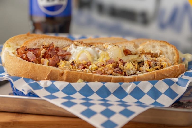 A loaded breakfast sandwich with bacon, eggs, cheese, and onions in a blue checkered basket.
