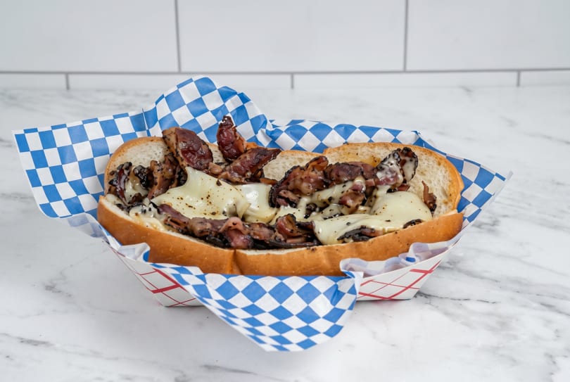 A cheesesteak sandwich with melted cheese and steak in a blue and white checkered paper tray.