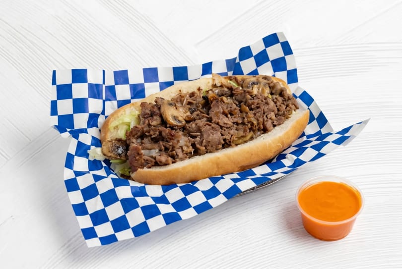 Cheesesteak sandwich with mushrooms and onions in a blue and white checkered paper, with a side of sauce.