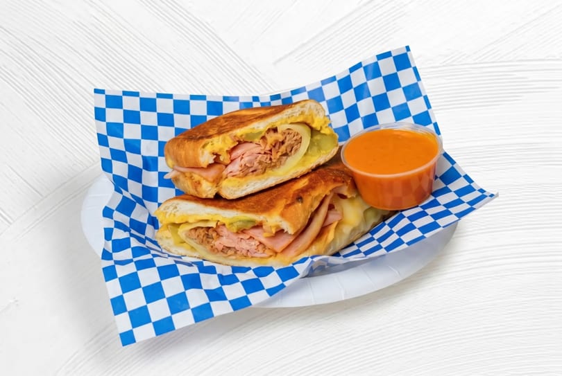 A Cuban sandwich cut in half, served on blue and white checkered paper with a side of orange sauce.