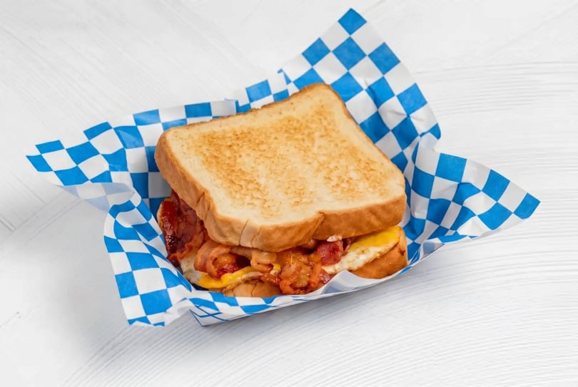 Breakfast sandwich with bacon, egg, and cheese on toasted bread, served in blue and white checkered paper.